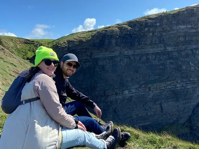 A couple sits on the grass at the edge of the Cliffs of Moher.