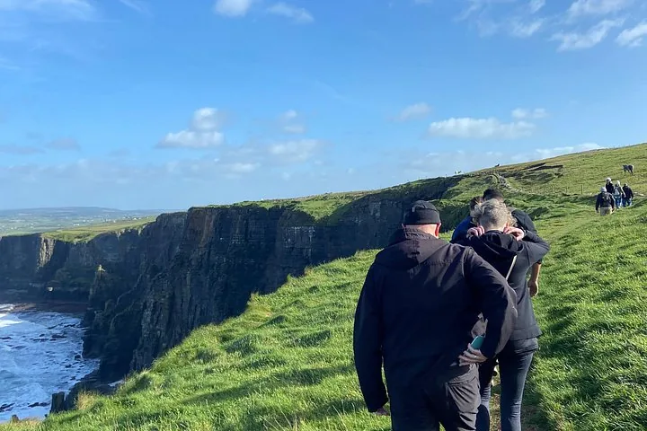 A group of people walk along a grassy path near the cliff edge.